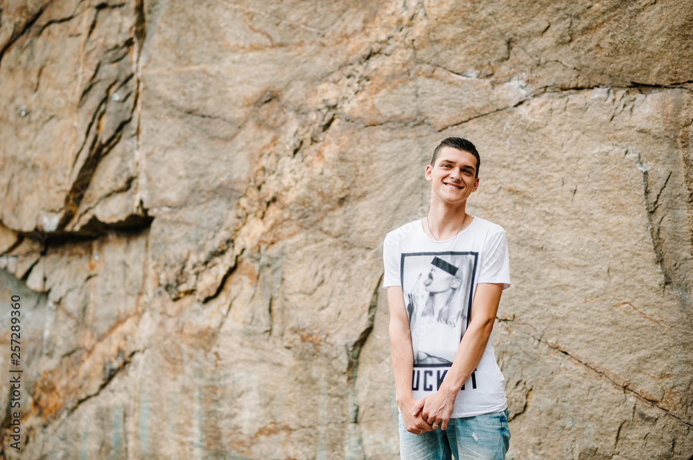 A man makes a grimace, stand on background of the large rocks wall on a big stone. upper half. looking at the camera. Close up. Place for text and design. Man poses at cliff of mountain on nature. A man makes a grimace, stand on background of the large rocks wall on a big stone. upper half. looking at the camera. Close up. Place for text and design. Man poses at cliff of mountain on nature.