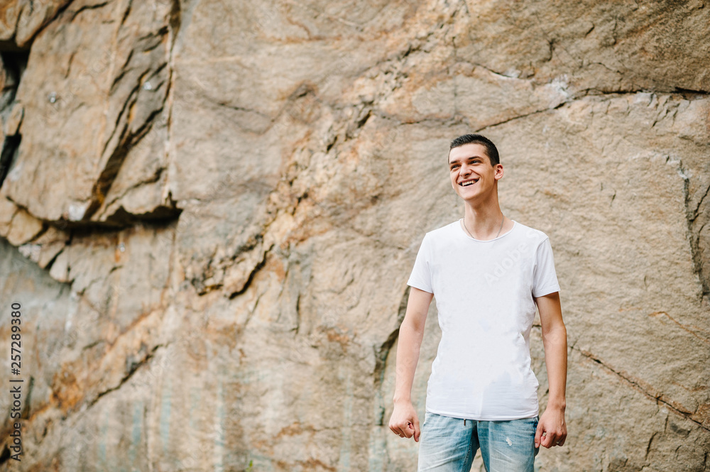 A man makes a grimace, stand on background of the large rocks wall on a big stone. upper half. looking at the camera. Close up. Place for text and design. Man poses at cliff of mountain on nature. A man makes a grimace, stand on background of the large rocks wall on a big stone. upper half. looking at the camera. Close up. Place for text and design. Man poses at cliff of mountain on nature.