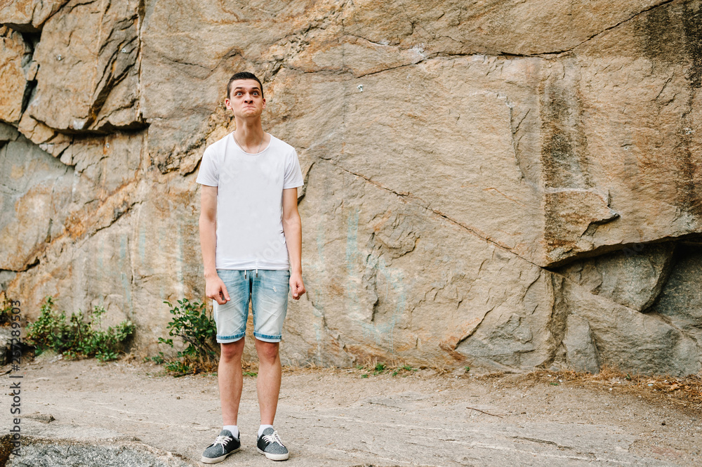 A man makes a grimace, stand on background of the large rocks wall on a big stone. full length. looking at the camera. Close up. Place for text and design. Man poses at cliff of mountain on nature. A man makes a grimace, stand on background of the large rocks wall on a big stone. full length. looking at the camera. Close up. Place for text and design. Man poses at cliff of mountain on nature.