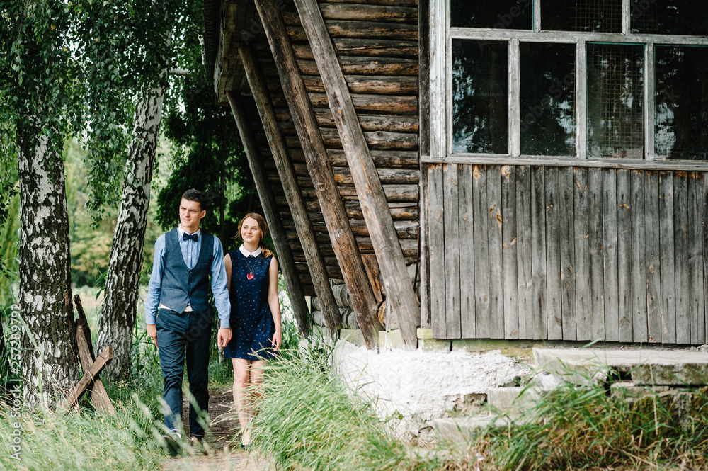 A young woman holds hand husband's, walking by birch tree near an old wooden stylish house. looking sideways. Young lover couple going on the background nature. full length. A young woman holds hand husband's, walking by birch tree near an old wooden stylish house. looking sideways. Young lover couple going on the background nature. full length.