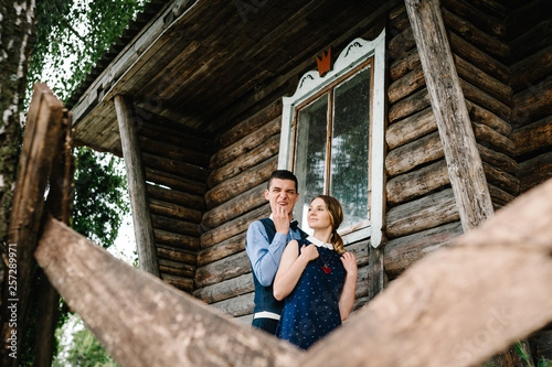 Man hugging woman from behind standing near old wooden stylish house. Young lover couple on the background nature. upper half length. Close up. The man frowns and make a grimace. © Serhii