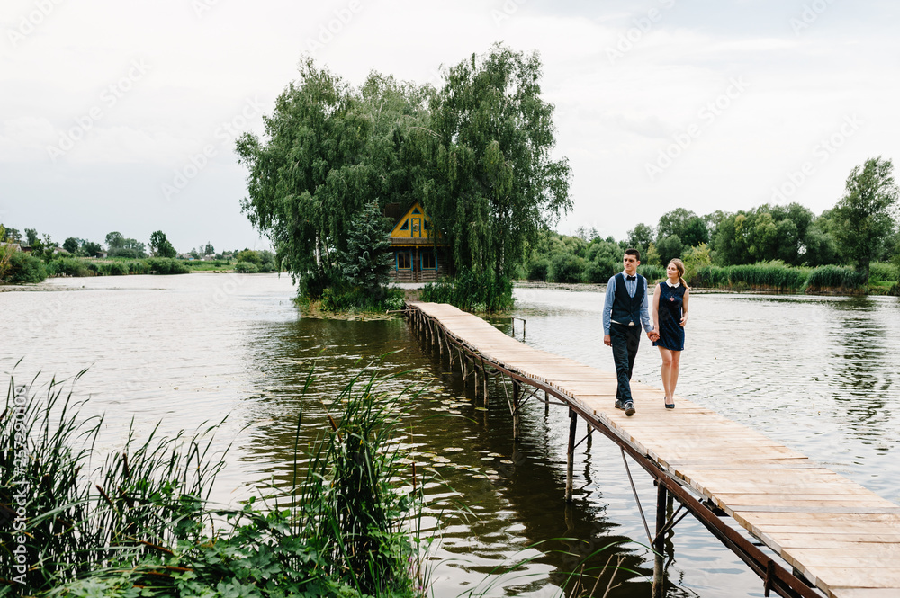 The young couple is holding hands, going on a wooden bridge above the lake on background a old house on an island near of trees. Nature, landscape. full length. The young couple is holding hands, going on a wooden bridge above the lake on background a old house on an island near of trees. Nature, landscape. full length.