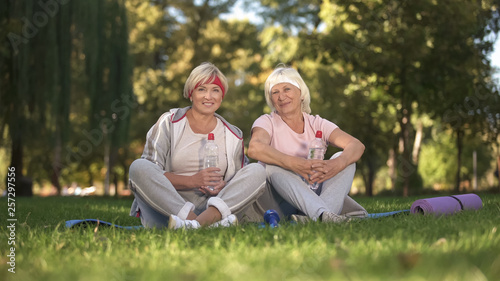 Two women sitting on grass and smiling into camera after finishing doing yoga