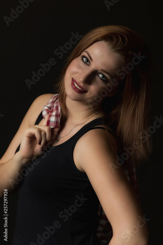 Low key portrait of happy young attractive woman on the black background. Woman is holding a checkered shirt over her right shoulder. Vertically. 