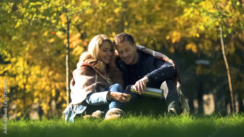 Fototapeta premium Man pouring hot tea from thermos, loving couple covered with plaid having picnic