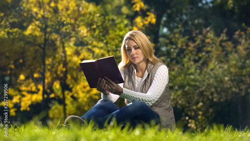 Concentrated woman reading scientific book in autumn park, sitting on grass © motortion