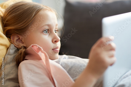Portrait of excited little girl using digital tablet while lying on sofa, watching cartoons or browsing internet, copy space
