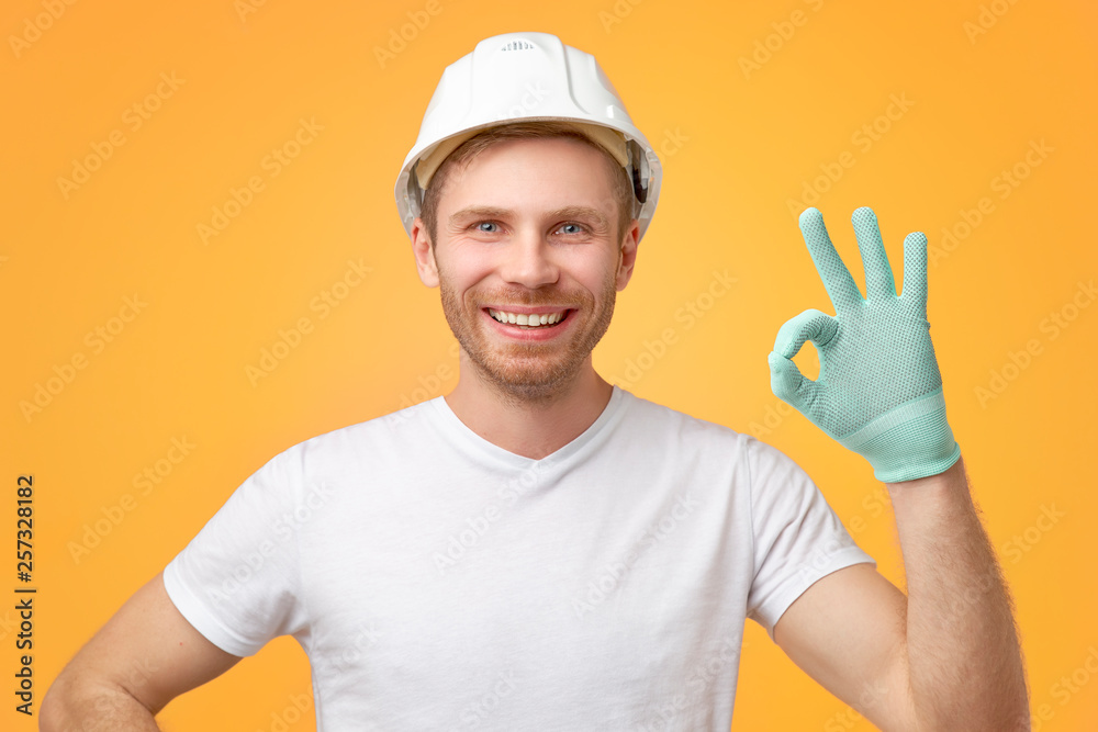 Pleased confident European man with broad smile, uniform, shows okay gesture, dressed in t-shirt and construction helmet. Isolated over white background