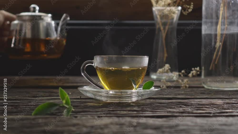 Hand holding jug and pouring hot organic tea into glass cup with tea leaves on wooden table with view of room on the morning, Vintage style Hand holding jug and pouring hot organic tea into glass cup with tea leaves on wooden table with view of room on the morning, Vintage style