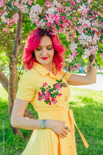 Beautiful and young woman with bright red hair and ref lips standing next to a blooming apple tree in a yellow dress. photo