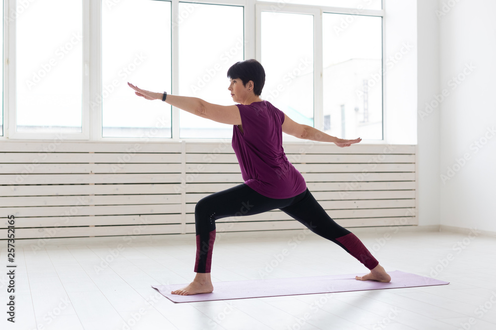 Fototapeta premium People, yoga, sport and healthcare concept - Middle-aged woman standing in Warrior over white background