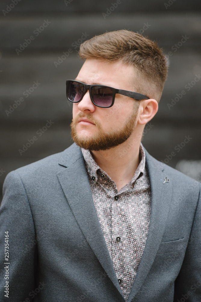 A man with a beard and sunglasses in a gray suit poses on the street to advertise men's clothing. Advertising menswear A man with a beard and sunglasses in a gray suit poses on the street to advertise men's clothing. Advertising menswear