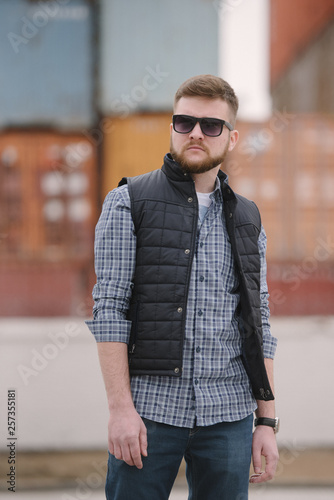 Slika na platnu A man with a beard and sunglasses in a plaid shirt and waistcoat posing on the street to advertise men's clothing