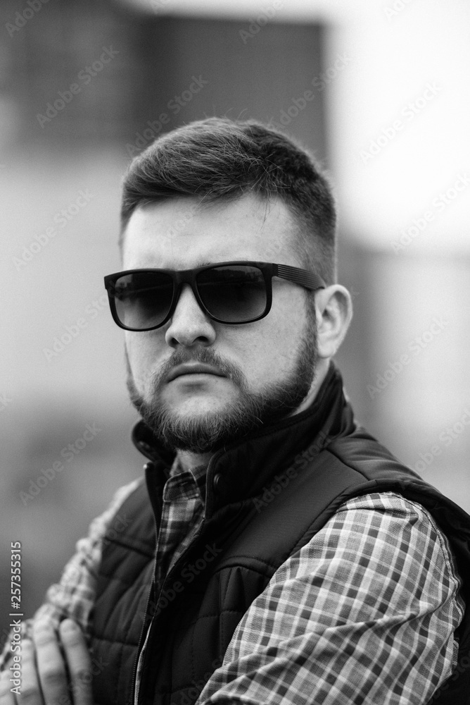 Obraz premium A man with a beard and sunglasses in a plaid shirt and waistcoat posing on the street to advertise men's clothing. Advertising menswear