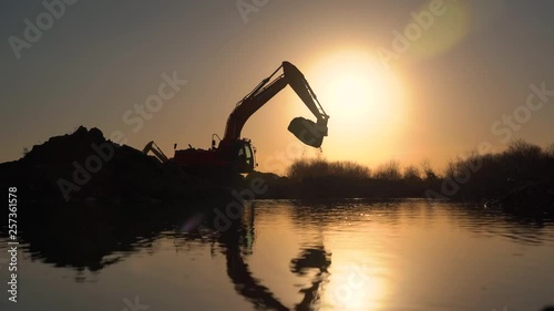 Silhouette of excavator. Dredge digs ground and loading tipper. Digger machine digging hole. Excavation of canal. Dump truck,tracked,bagger,backhoe,shovel,sunny blue sky,sun in background,backlight.