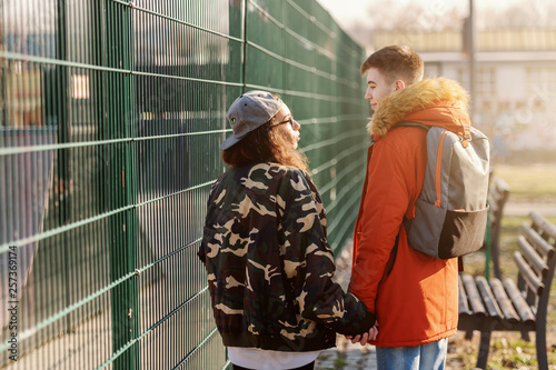 Cute teenage couple holding hands and walking next to fence. Backs turned.
