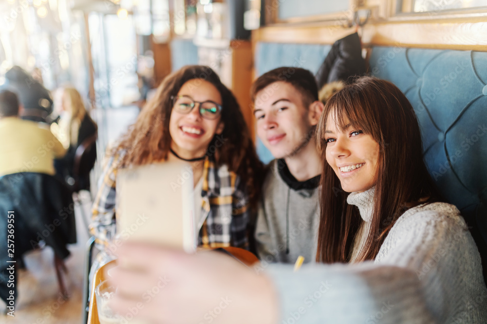 Fototapeta premium Three multicultural teenage classmates taking selfie while sitting at cafeteria.