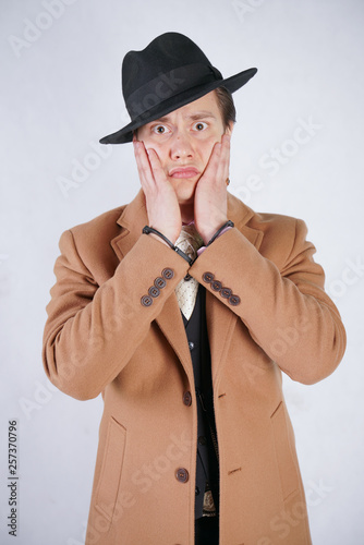 young man in a black business suit and beige fashion coat with a stylish hat stands in police handcuffs on a white solid Studio background
