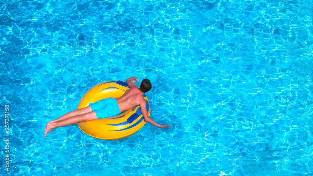 Young man is floating on yellow inflatable air ring/circle in pool with blue water. Holiday leisure in happy sunny day. Vacation concept, top view.
