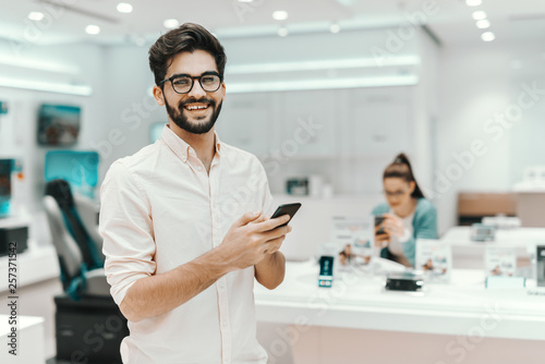 Attractive mixed race man with eyeglasses holding smart phone while standing in tech store.