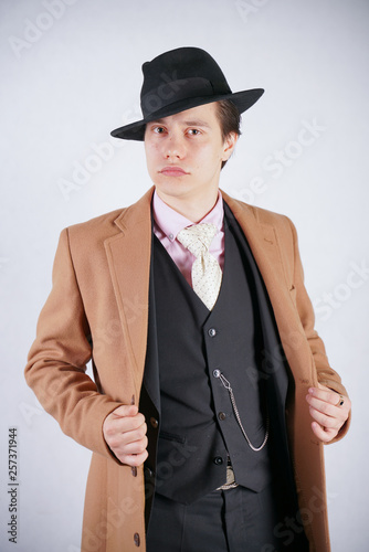 fashionable young man in black business suit and beige coat with hat on white studio background alone