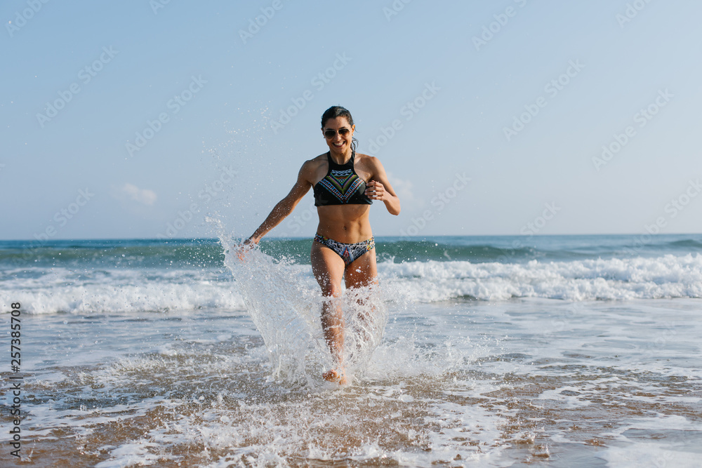 Fit young woman in bikini running at the beach into the sea splashing water drops. Outdoor fitness, healthy lifestyle and summer freedom concept. Fit young woman in bikini running at the beach into the sea splashing water drops. Outdoor fitness, healthy lifestyle and summer freedom concept.
