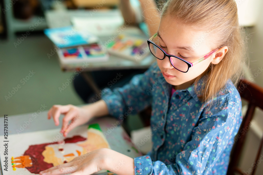 Fototapeta premium Portrait of adorable little girl smiling happily while enjoying art and craft lesson in art school working together with other kids