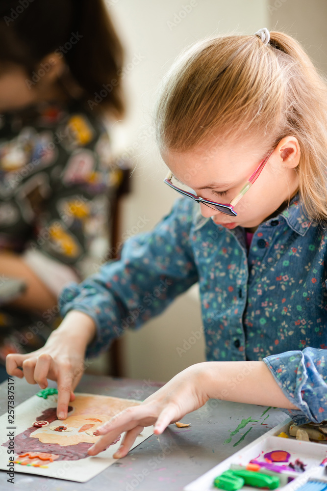 Portrait of blonde girl with big black glasses smiling happily while enjoying art and craft lesson in art school working together with other kids