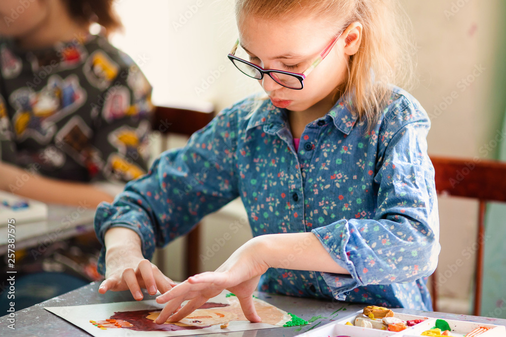 Fototapeta premium Portrait of blonde girl with big black glasses smiling happily while enjoying art and craft lesson in art school working together with other kids