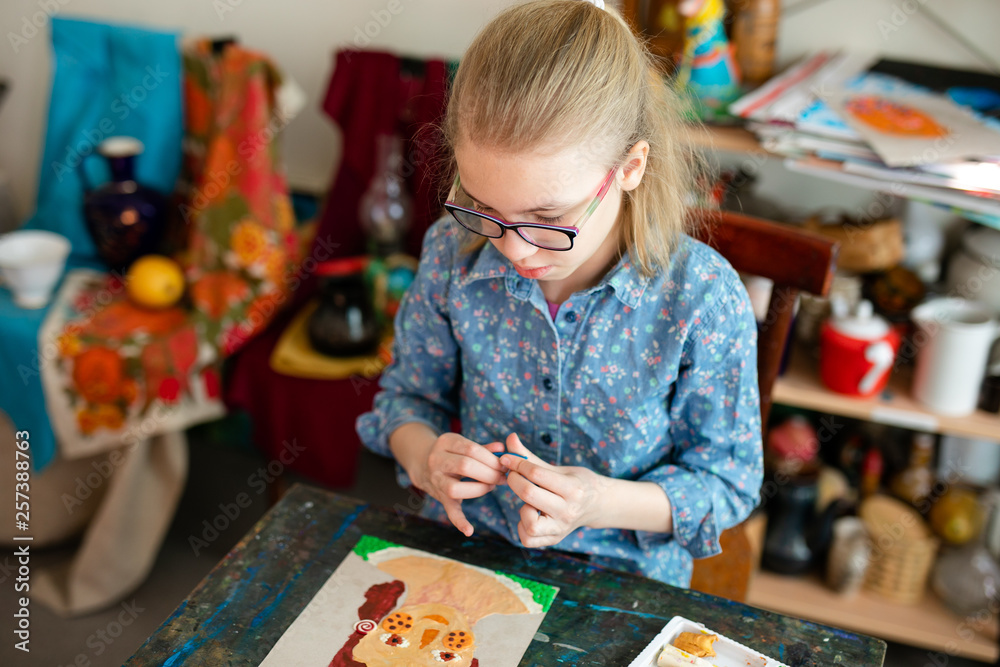 Portrait of blonde girl with big black glasses smiling happily while enjoying art and craft lesson in art school working together with other kids