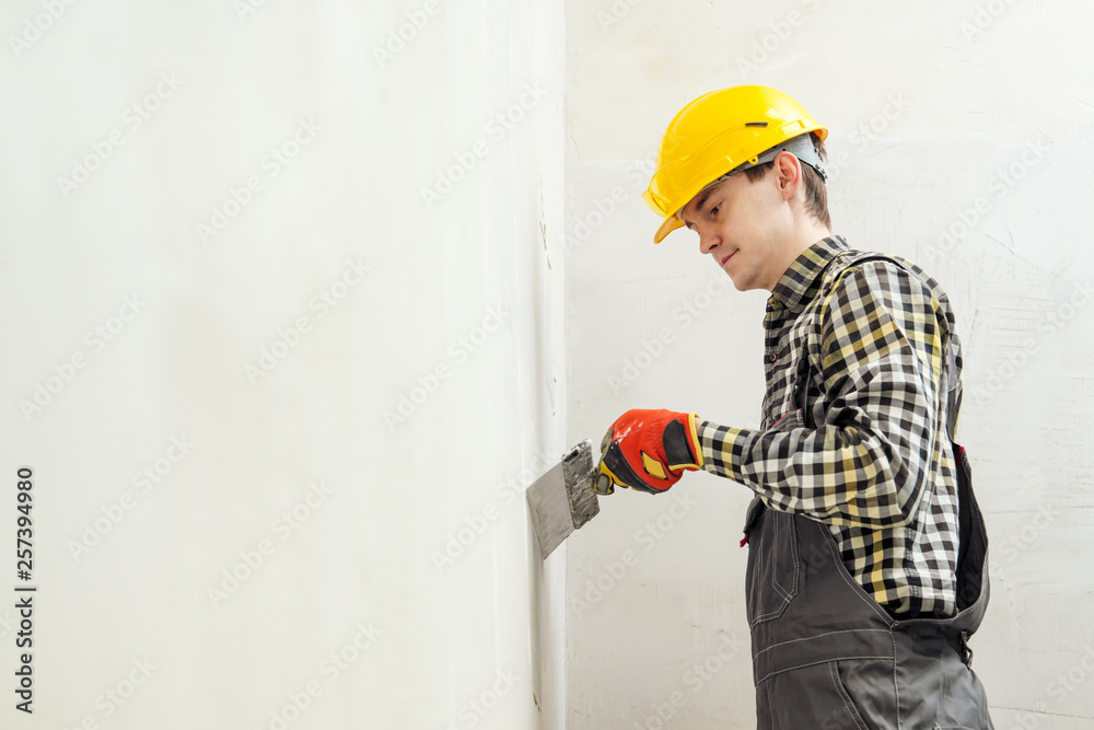 Young male builder and repairman in a yellow helmet plastering the wall with a spatula and plaster.