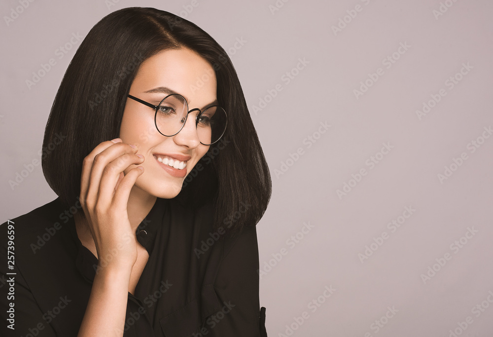 Portrait of happy smiling young brunette businesswoman in glasses, over grey background, with copyspace area for slogan or text message. Model in black shirt and glasses. Office management, eye care Portrait of happy smiling young brunette businesswoman in glasses, over grey background, with copyspace area for slogan or text message. Model in black shirt and glasses. Office management, eye care