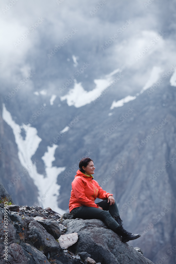 Girl smiling in a red jacket sitting on the mountain. Enjoys the beautiful view around.
