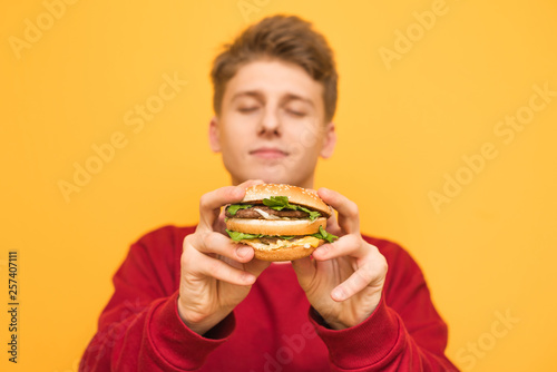 Handsome guy with his eyes closed shows a delicious large burger to the camera  holds in his hands  isolated on a yellow background.Focus on an appetizing burger in the hands of a young man. Fast food