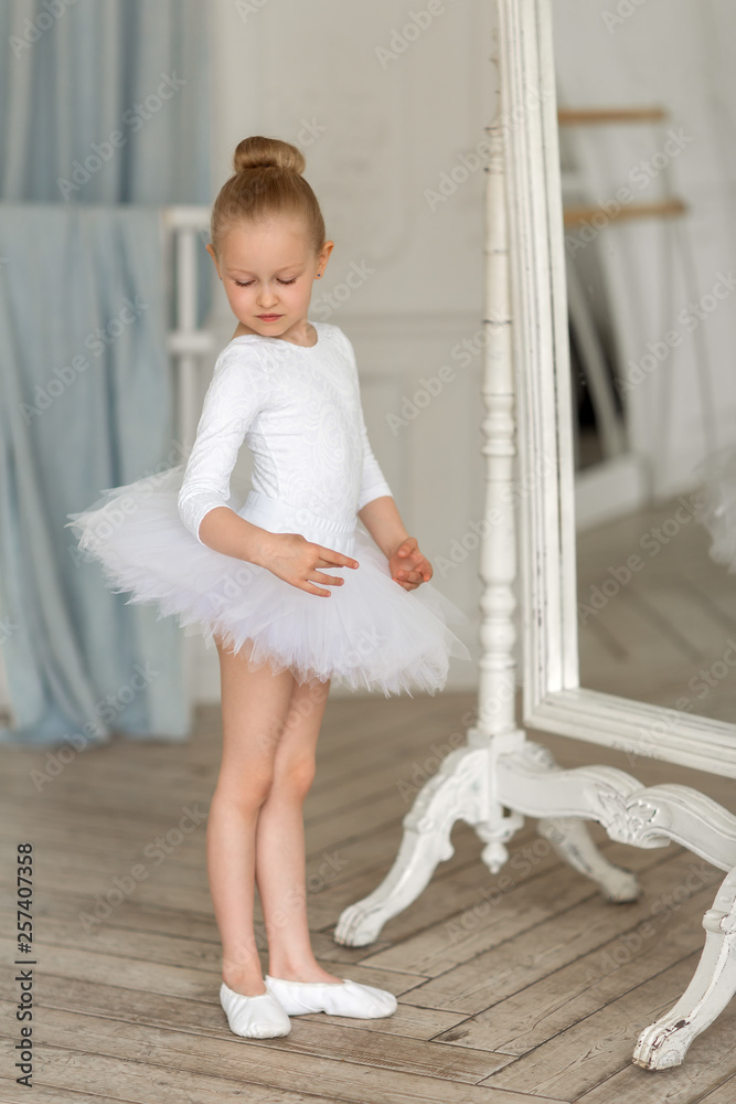 Foto Stock A little sweet ballerina in a white tutu stands by the mirror  and admires herself. The Ballet. | Adobe Stock