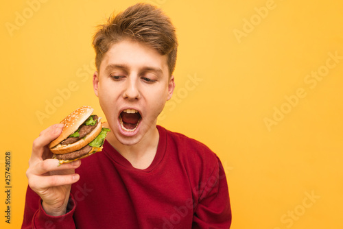 Portrait of an emotional young man with a burger in his hands on a yellow background. A hungry emotional guy holds a burger in his hands and shouts  isolated. Very hungry student. Copyspace