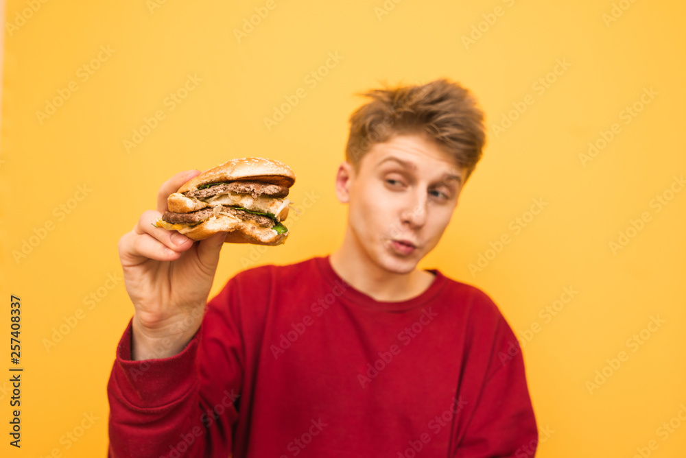 Handsome young man in the casual clothing holds an oozing burger in his hand and looks at him, isolated on a yellow background. Appetizing burger in the hands of a guy. Handsome young man in the casual clothing holds an oozing burger in his hand and looks at him, isolated on a yellow background. Appetizing burger in the hands of a guy.