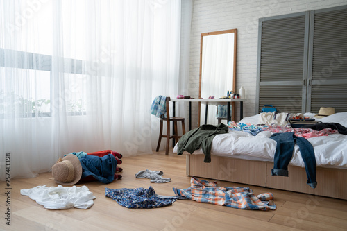 straw hat and colorful clothes in luggage on wooden floor. empty nobody in messy white bed in bedroom packing suitcase for travel abroad summer vacation holidays. mirror by window at dressing table. photo