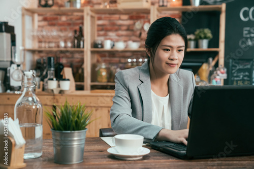 Attractive asian female office worker is typing messages on laptop computer while sitting in coffee shop. businesswoman insurance sales working on portable pc in cafe. elegant lady in suit smile