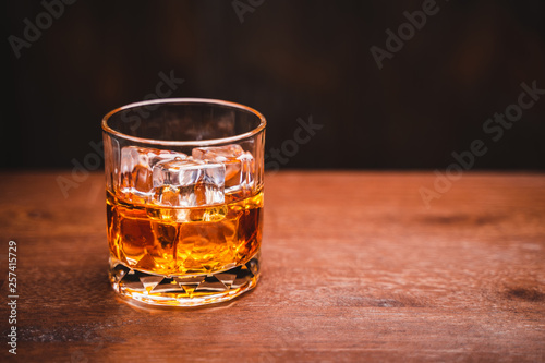 Glass of whiskey with ice cube on wooden table over black background