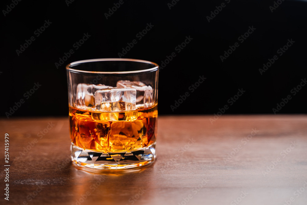 Glass of whiskey with ice cube on wooden table over black background Glass of whiskey with ice cube on wooden table over black background