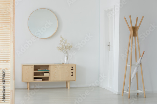 Wooden hanger with white umbrella in real photo of entrance hall interior with cupboard with books and plant in glass vase and round mirror on the wall