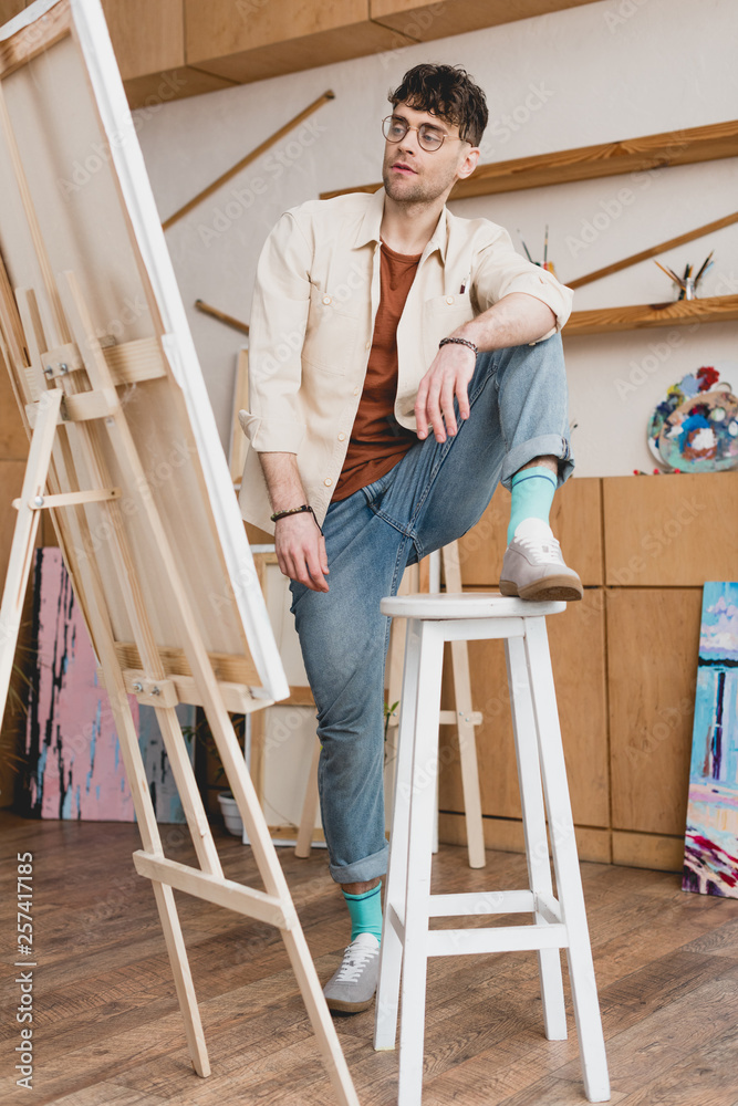handsome artist standing with foot on chair and looking at easel with canvas