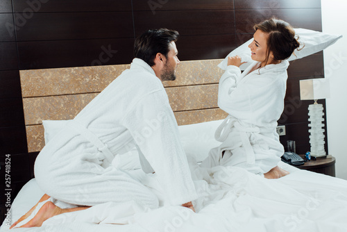 happy couple in white bathrobes having pillow fight on bed