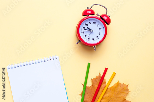 Alarm clock, empty notebook, pencils on yellow background.