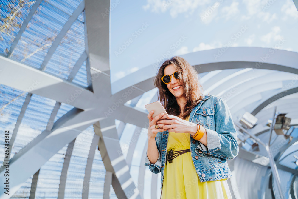 Fototapeta premium Woman wearing yellow shirt texting on the smart phone walking in the street in a sunny day