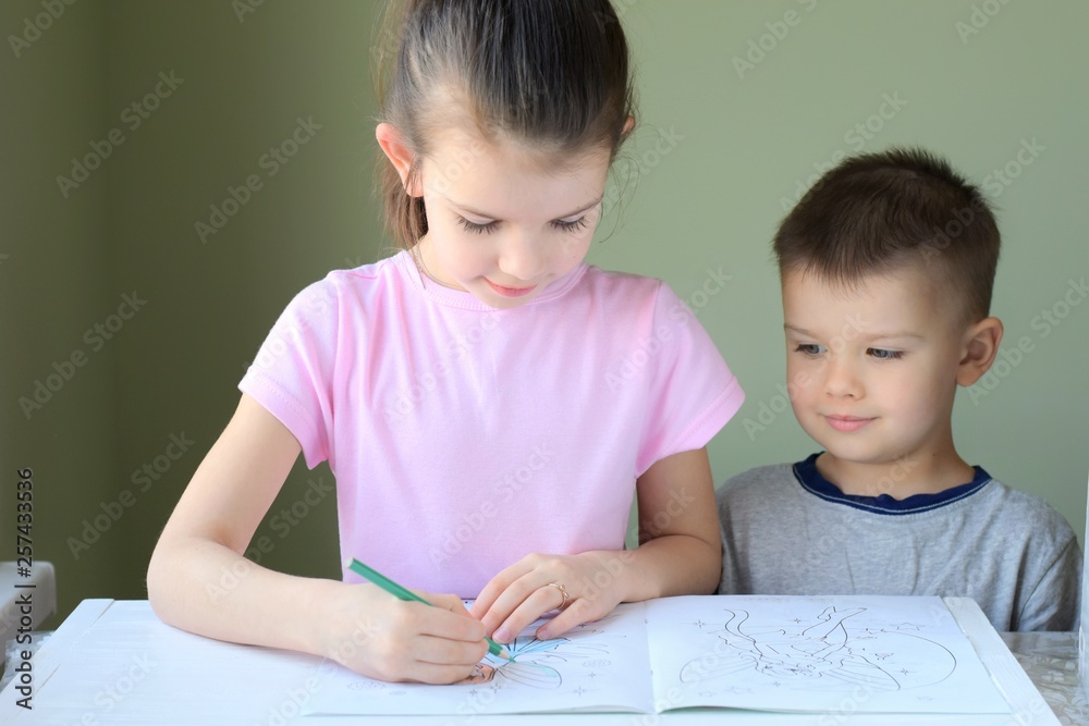 Little cute boy and girl draw together with colorful pencils on blurred neutral background. Beautiful white caucasian girl learns to draw her little toddler brother. Smiling children learning art Little cute boy and girl draw together with colorful pencils on blurred neutral background. Beautiful white caucasian girl learns to draw her little toddler brother. Smiling children learning art