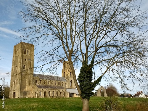 Landscape view of ancient Wymondham Abbey, historical site in Norfolk East Anglia England recently refurbished with towers from across river with green grass lawn and trees blue skies on Spring day photo