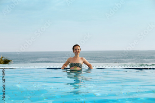 Luxury Resort. Woman Relaxing In Infinity Swimming Pool Water. Beautiful Happy Healthy Female Model Enjoying Summer Travel Vacation  Looking At Sea View. Summertime Recreation  Relax And Spa Concept.
