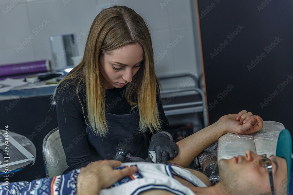 A beautiful blonde girl tattooes the arm of a boy in her tattoo studio
